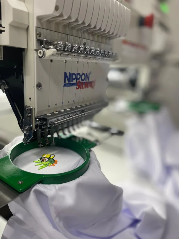 Máquina de bordar industrial aplicando um bordado colorido em tecido preso a um bastidor verde